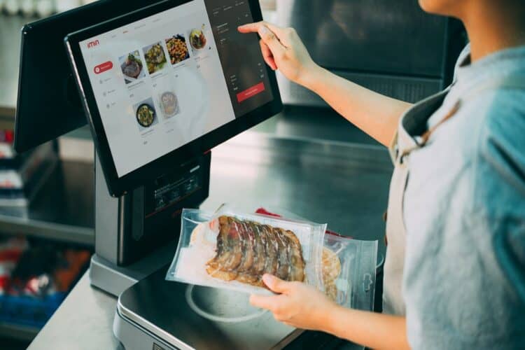Photo by iMin Technology: https://www.pexels.com/photo/person-standing-in-front-of-a-monitor-holding-a-pack-of-food-and-12935041/