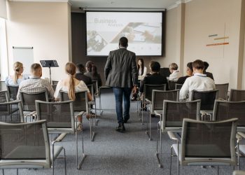 Photo by Pavel Danilyuk from Pexels: https://www.pexels.com/photo/a-man-walking-on-aisle-between-chairs-with-people-sitting-8761317/